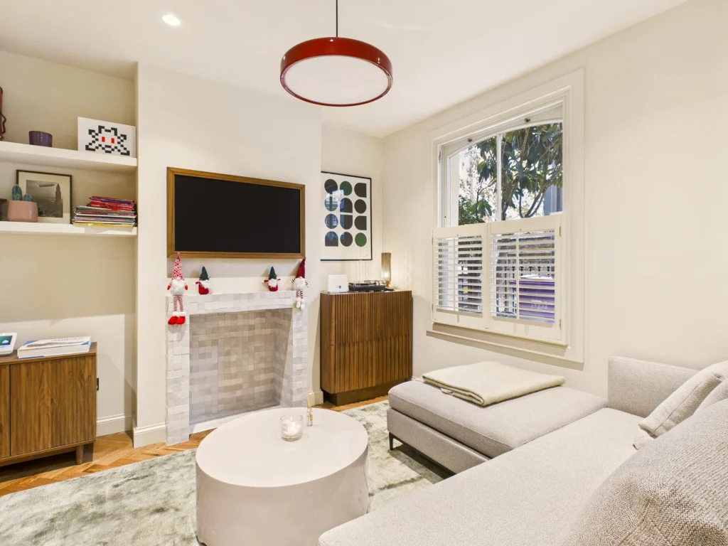 Snug living room with a white tiled fireplace, walnut cabinets, a red pendant light and a sash window with shutters