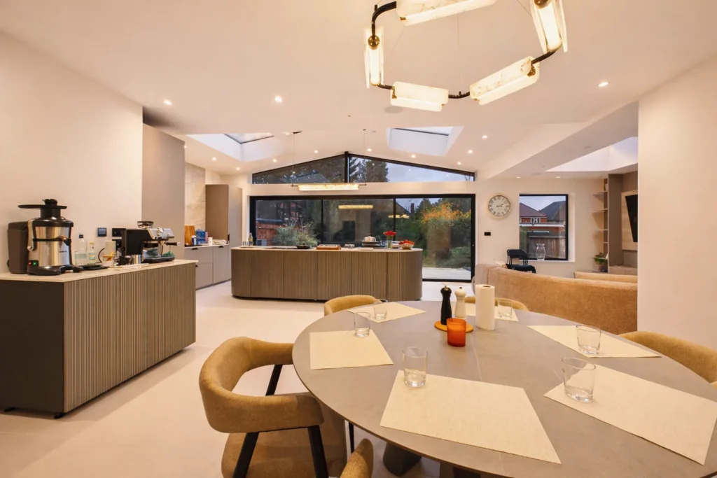 Open-plan kitchen and dining area with a round dining table, an alabaster ring pendant, fluted kitchen units, rooflights and full-width sliding doors to the garden