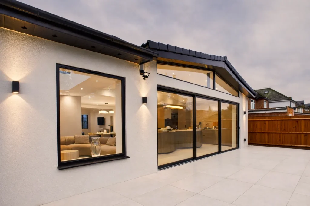 Rear wraparound extension at dusk, with a large picture window to the living area, black sliding doors to the kitchen and a porcelain terrace