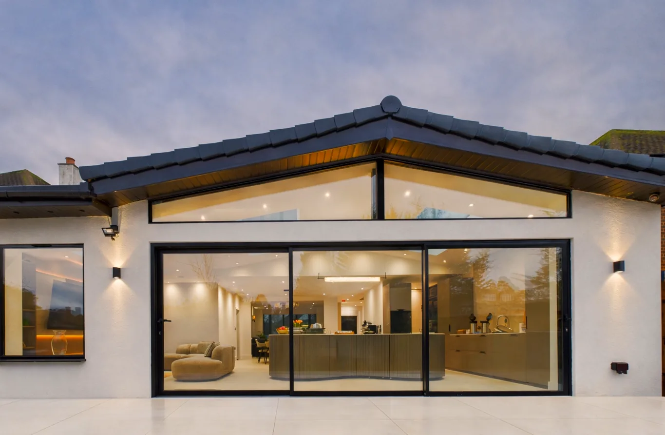 Rear wraparound extension with a dark tiled pitched roof, triangular clerestory glazing and full-width black sliding doors onto a pale porcelain terrace