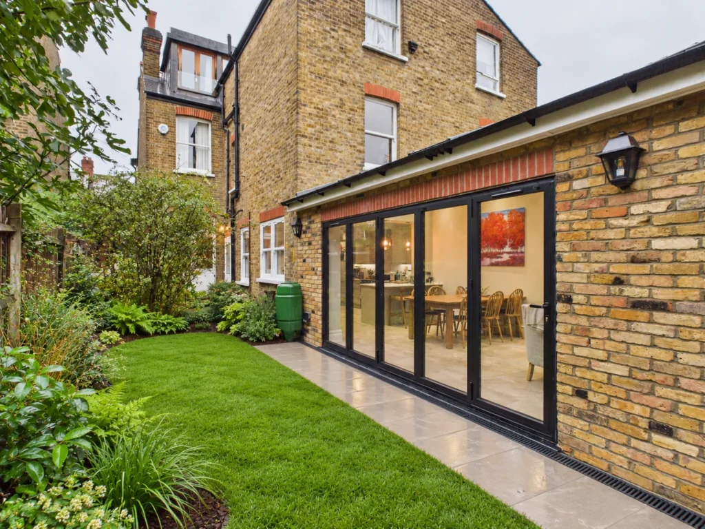 Single-storey rear extension in London stock brick with black aluminium bifold doors along the flank, opening onto porcelain paving and a lawn