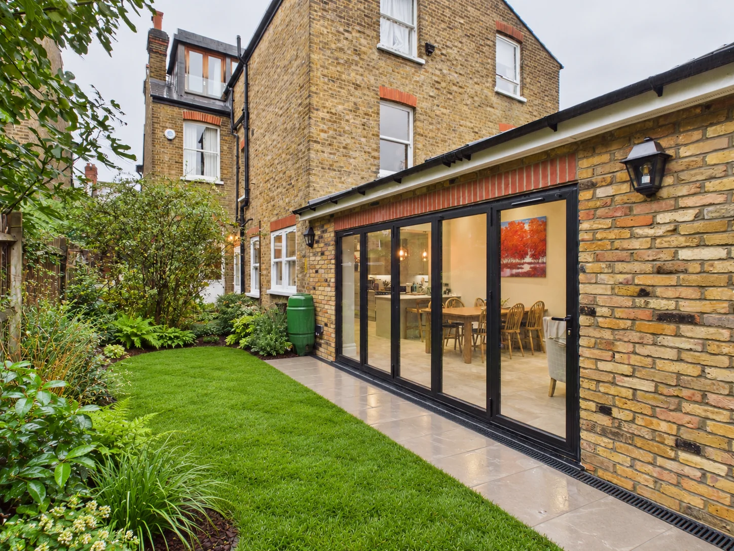 Single-storey rear extension in London stock brick with black aluminium bifold doors along the flank, opening onto porcelain paving and a lawn