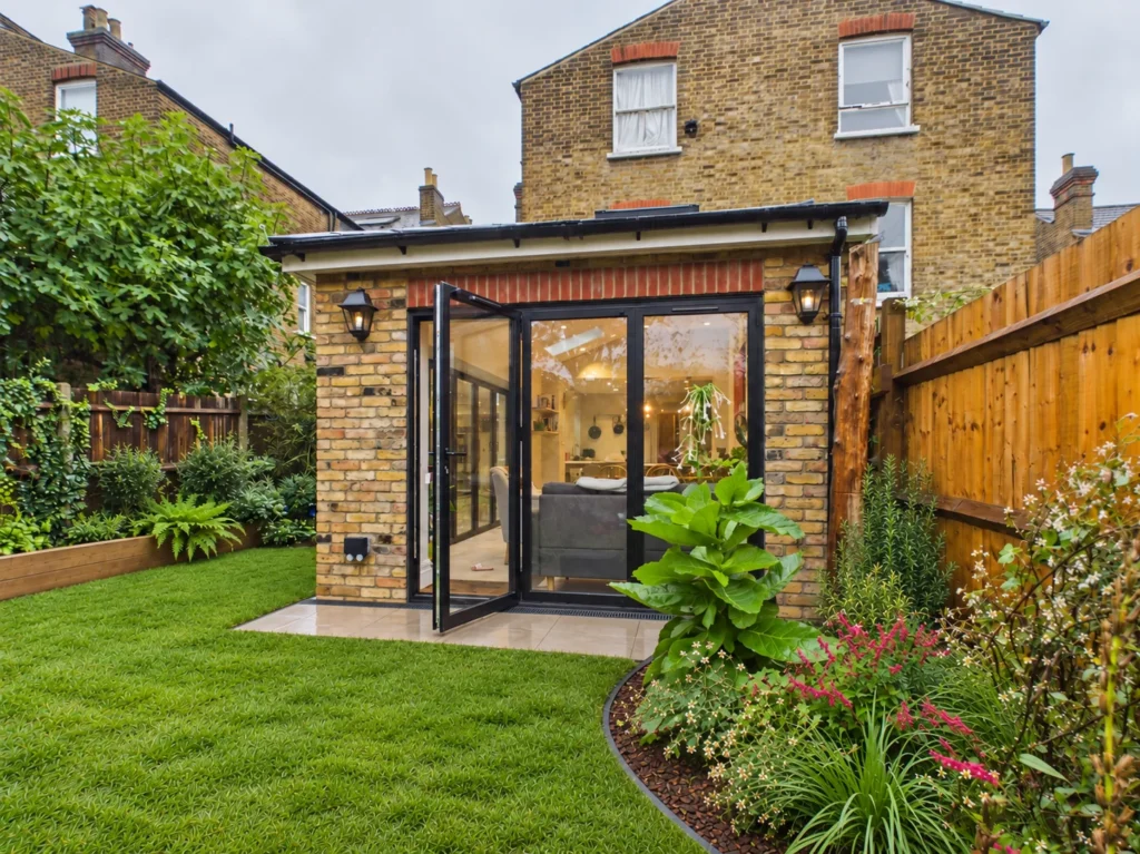 Rear elevation of a single-storey London stock brick extension with black aluminium bifold doors, coach lanterns and a red brick soldier course, facing a lawn and planted border